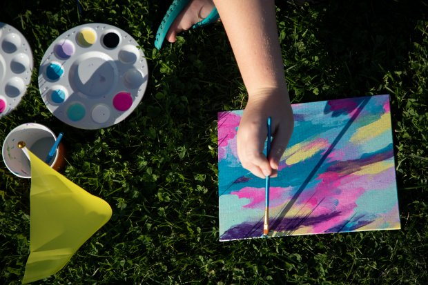 A student paints abstract art on a small canvas while seated in the grass during the Student Association's Paint & Vibe Event