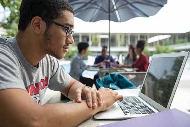 A male student uses a laptop while seated outside on campus.
