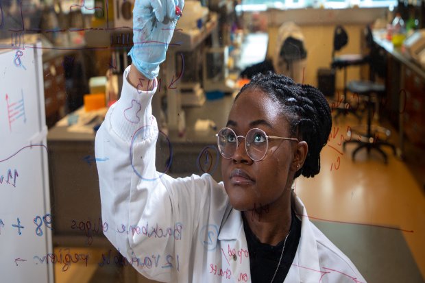 A woman with braids wearing a white lab coat, blue gloves and wire frame glasses writes in erasable marker on a clear board.