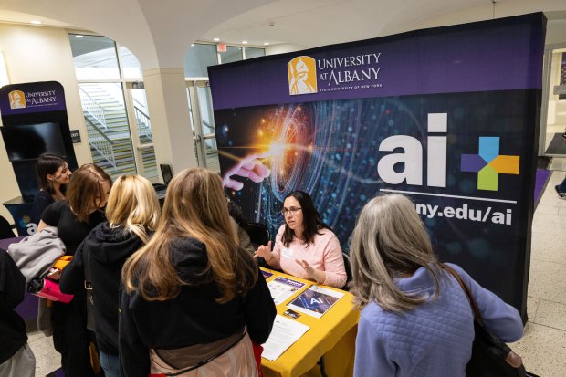 A UAlbany staff member sits at an information table with a large AI Plus sign and speaks to gathered students, faculty and staff.