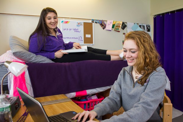 Two students, one sitting a bed with a textbook and the other sitting at a desk with a laptop, laugh inside a residence hall room