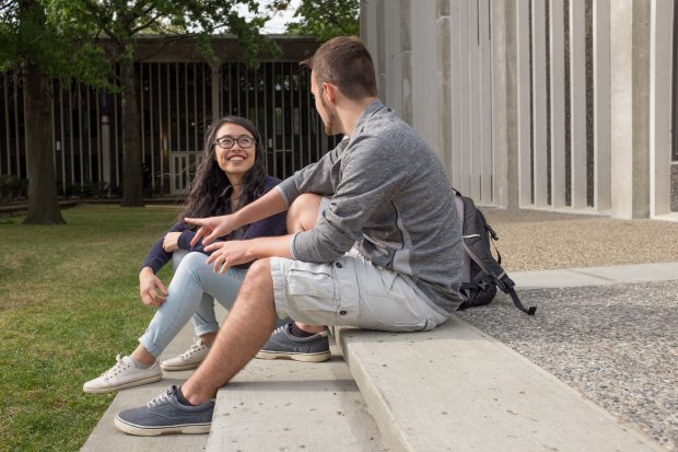 Two students sit on the steps outside a residence hall, laughing and talking in the sun