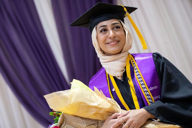A student wearing a hijab, graduation cap, gown and sash smiles for a photo while holding flowers.