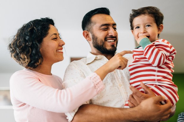 ponytail.  A Latina mother in a pink blouse and a Latino father in a white short-sleeved button up shirt smile at their young son. The father is holding the son, who is smiling with his hand in his mouth while wearing red and white striped pajamas.