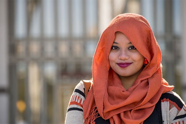 A UAlbany student wearing burnt orange hijab and white and orange sweater smiles for a portrait on uptown campus.