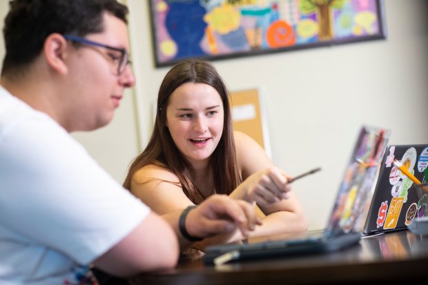 A Writing Center peer tutor smiles and points something out on another student's laptop screen.