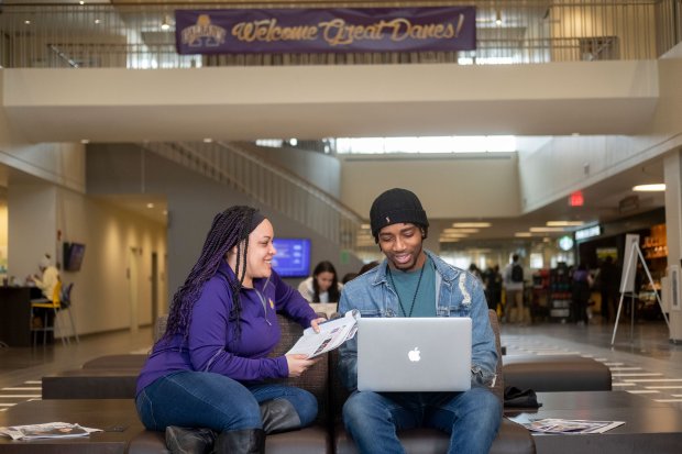 Two students sit inside the Great Dane Student Union, smiling and holding notebooks and laptops.