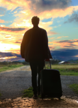 A man stands silhouetted against a vibrant sunset. He is pulling a roll-along suitcase.