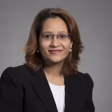 A woman in glasses, a black blazer and white blouse with shoulder-length brown hair poses for a portrait against a gray backdrop