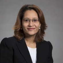 A woman in glasses, a black blazer and white blouse with shoulder-length brown hair poses for a portrait against a gray backdrop