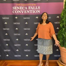 A woman with dark hair and glasses, wearing an orange blouse and gray skirt stands in front of a backdrop that reads, "175th Anniversary of the Seneca Falls Convention," with repeating NYS logos.
