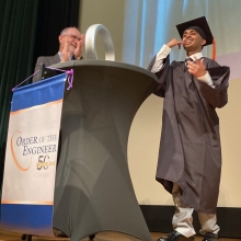New engineering graduate poses on stage with his ring while department chair applauds.