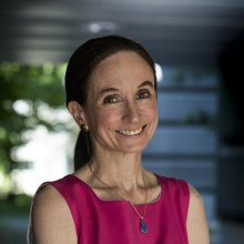 Kathryn Hess smiles, with sun and greenery in the backgroud, wearing a sleeveless red top, a necklace and her hair pulled back in a ponytail.