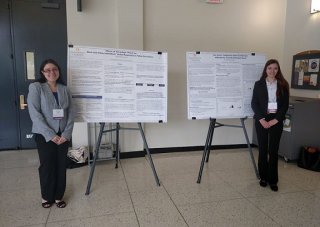 Two women, dressed in business apparel and wearing name tags smile and stand on either side of two research posters displayed side-by-side
