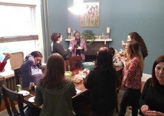 Eleven people sit and stand around a dining room table, smiling, talking and enjoying food.