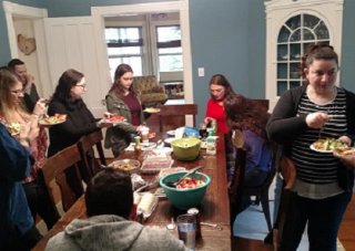 Ten people sit and stand around a dining room table, eating, talking and laughing