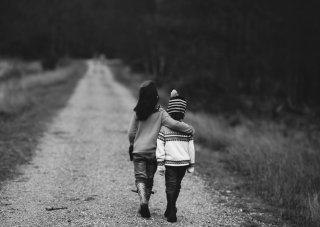 Two young children walk together down a wooded path; the taller child has their arm around the smaller one. 