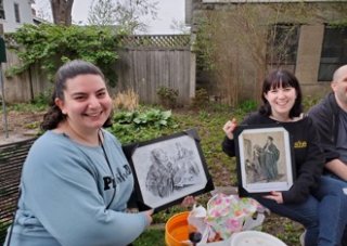 Two women with brown hair sit in a backyard, smiling while holding up framed historical drawings.