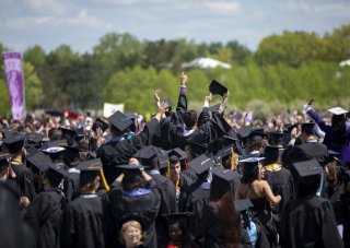 Una multitud de graduados de UAlbany con túnicas negras de graduados y gorras de mortero celebrando la graduación con los brazos levantados en el aire.