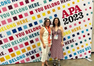 Two women wearing lanyards stand in front of a colorful pop-up wall that reads, 'you belong here.' and 'APA 2023'