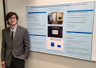 A man with brown hair, a pale blue shirt, and a gray blazer stands to the left of a research poster titled, 'Do 'Groove' Inducing Sounds Need to be Perceived as Music for Individuals to Show Movement?'