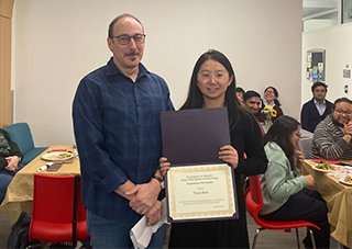 A woman in a black dress with long, dark hair holds up a certificate and smiles as she stands on the right side of the image, next to man in a blue plaid button up and glasses.