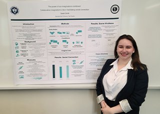 A woman with long brown hair, a white blouse and a black blazer smiles while standing next to a research poster titled, 'The power of our imagination combined: Collaborative imagination's role in facilitating social connection,' with a byline that reads, 'Sarah Smith.'