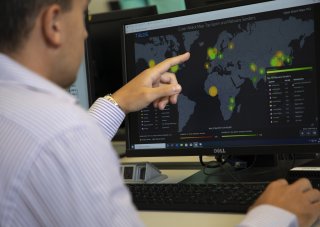 A man in a button-down shirt points at a computer displaying a world map and glowing with yellow and green circles. The map says "Cyber Attack Map Top Spam and Malware Senders" at the top.