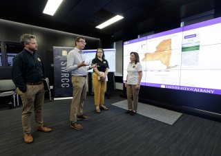 Governor Kathy Hochul stands with Nick Bassill and other New York emergency management leaders in front of a weather map inside the xCITE lab.