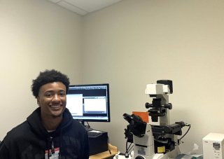 A smiling young man wearing a black jacket and clip on name badge sits in front of a computer screen next to a microscope.