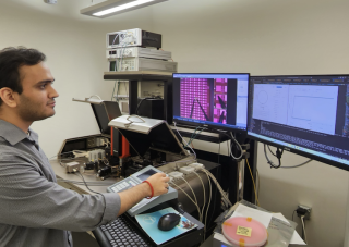 A man with short dark hair in a gray shirt stands in front of two computer screens in a laboratory.