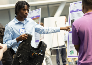 A man with short black braids in a light blue shirt holds up a black vest and gestures to a poster as he speaks to a man standing nearby in a purple shirt