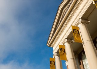 A bright blue sky accentuates the roof of a brick building with ornate columns holding yellow UAlbany flags