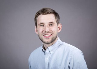 A man wearing a light blue button-down shirt smiles for a portrait against a gray backdrop.