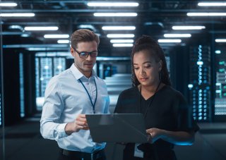 Two cybersecurity professionals standing in a server room consult a laptop.
