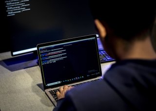 A man sits at a desk with a desktop computer and types code on a laptop.
