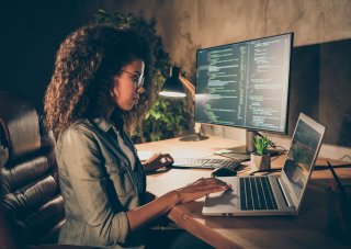 A cybersecurity professional works on a laptop alongside a monitor displaying code.