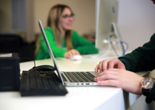 Two hands work at a laptop upon a white desk in the foreground, while a woman in a green shirt and long hair works at a computer in  the background, out of the camera's focus.
