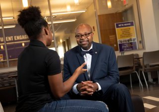 Dean of Students Clarence L. McNeill smiles as he sits outside his office speaking with a student, who is faced away from the camera.