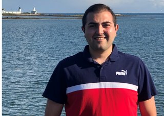 Bradey Liverio stands in front of body of water, wearing a navy, white, and red collared shirt