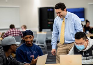 An instructor in a blue button up and yellow tie stands and speaks with three students seated around a table.