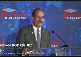 A screenshot of President Rodríguez standing alone at a podium in a gray suit and addressing the audience at the Seal of Excelencia announcement. 
