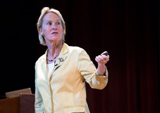 Frances Arnold stands onstage wearing a yellow blazer and holding a powerpoint pointer in her left hand.