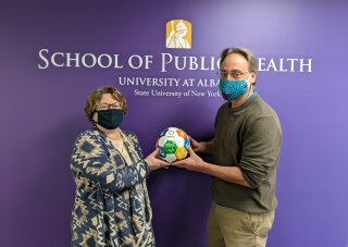 John and Lynne stand in front of a purple wall that says "The School of Public Health" in white text. They are wearing face masks and are holding a soccer ball from the UN that has the Sustainability Goals written on it.