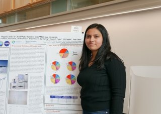 A woman with long dark hair wearing a long-sleeved black shirt stands in front of a presentation poster.