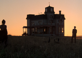 Still from Terrence Malick’s film Days of Heaven (1978), includes house and landscape in shadows with two people standing on either side.