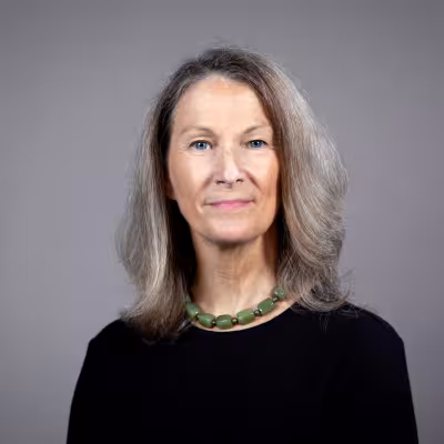 Margi Sheehy in black top and green neckace with shoulder length hair, gray background