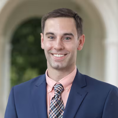 Dr. Jonathan Foster smiling blue suit jacket plaid tie peach shirt campus arch background.
