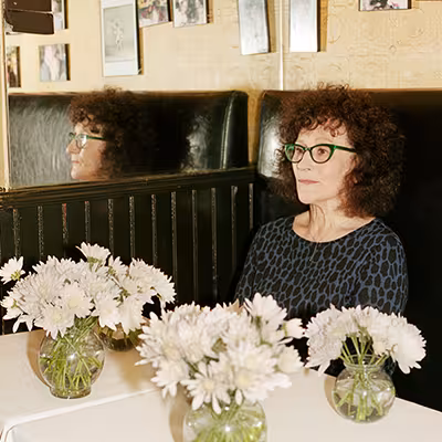 Lynn Tillman sits in a booth at a restaurant in a gray patterned blouse and glasses. Three vases with while flowers sit on a table before her.