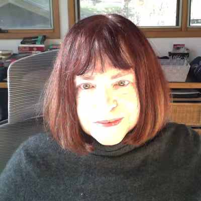 Mary Valentis sits at her desk. She wears a gray sweater and her auburn hair is cut shoulder-length with bangs.